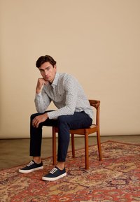 Witte bloemenbedrukte overhemd met blauwe accenten, donkerblauwe broek, marineblauwe sneakers met witte zolen, zittend op een houten stoel op een geweven tapijt.