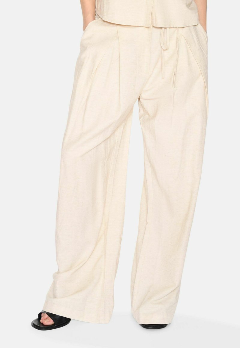 Weite Hose aus einem leichten beigen Stoff mit einer weichen Textur, ausgestattet mit Falten und einem verstellbaren Kordelzugbund. Minimalistisches Design.
