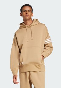 Sudadera color arena con bolsillo frontal, cordones y tres rayas blancas en una manga. Presenta un pequeño logo blanco en el dobladillo. Tejido suave.