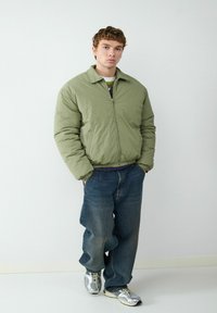 Jasnozielona kurtka puchowa z zamkiem z przodu, połączona z luźnymi niebieskimi dżinsami i srebrnymi trampkami. Gładka tekstura, swobodny krój.