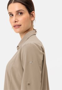 Camicia beige con colletto, caratterizzata da una texture liscia, con un logo ricamato discreto sulla manica sinistra e dettagli con bottoni sui polsini.