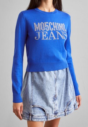 Kobieta ubrana w niebieski sweter z długim rękawem z napisem "MOSCHINO JEANS" z kryształków oraz jasną spódnicę dżinsową w kształcie litery A z przednimi kieszeniami.