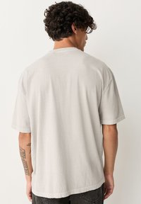 Maglietta a maniche corte di colore grigio chiaro, realizzata in tessuto morbido, con vestibilità comoda e design semplice. Tatuaggio visibile sul braccio sinistro.