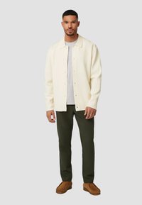 Lichte crèmekleurige button-up jas over een grijze shirt, gecombineerd met donkergroene broek en bruine schoenen, staande tegen een neutrale achtergrond.