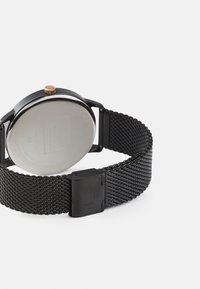 Black mesh strap watch with a round black case and a rose gold crown. Features a smooth, reflective silver back and secure clasp design.