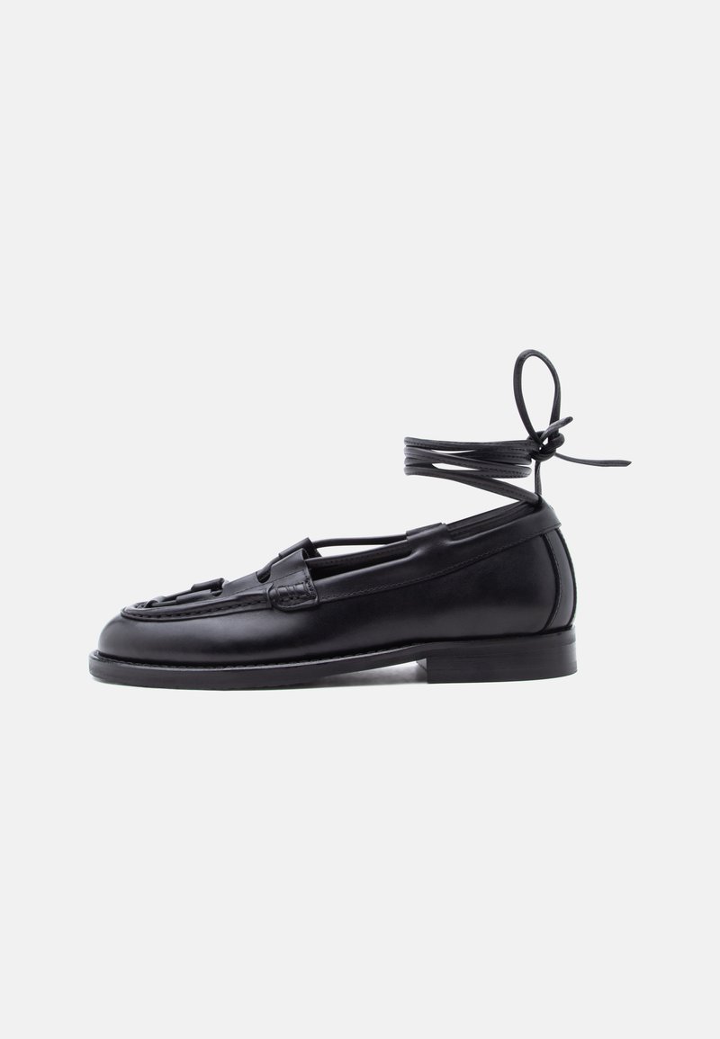 Mocassin en cuir noir avec semelle plate et lacet autour de la cheville noué en un nœud, présenté sur un fond clair uni.