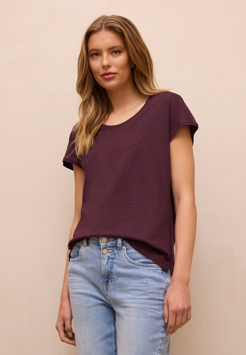 Jeune femme aux longs cheveux blonds portant un t-shirt violet foncé à manches courtes et un jean bleu clair sur un fond beige uni.