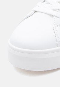 Λευκό αθλητικό παπούτσι  με λεία συνθετική επάνω επιφάνεια, στρογγυλεμένη μύτη, επίπεδα κορδόνια και παχιά λευκή σόλα με διακριτική υφή. Απλός σχεδιασμός, χωρίς σχέδια.