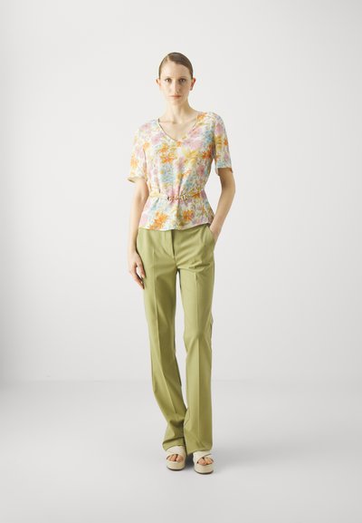 Bluse mit Blumenmuster in Pastellfarben, V-Ausschnitt und kurzen Ärmeln, kombiniert mit hellgrünen, geraden Hosen und beigen Sandalen.