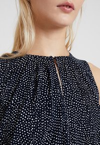 Marinblus med vita prickar, med en nyckelhålsklänning och veckad design. Tillverkad av mjukt tyg.