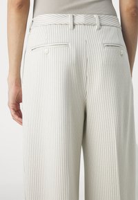 Pantalon blanc avec de fines rayures verticales noires, poches arrière avec boutons, porté par une personne avec un haut côtelé beige.