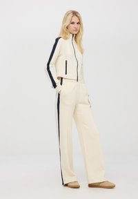 Giacca beige con zip, strisce laterali blu navy e due tasche con zip, abbinata a pantaloni beige coordinati con dettagli blu navy. Tessuto morbido.