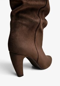 Botte à talon en daim marron avec une texture lisse, un design plissé et un talon effilé. Présente des coutures visibles le long du côté.