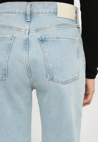 Light blue denim jeans with a mid-rise waist, featuring two back pockets and a stitched seam, complemented by a tan leather tag at the waistband.