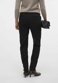 Pantalons noirs ajustés avec une coupe slim, dotés de poches arrière. Associés à un haut beige et tenant une pochette noire, portés avec des talons bordeaux.