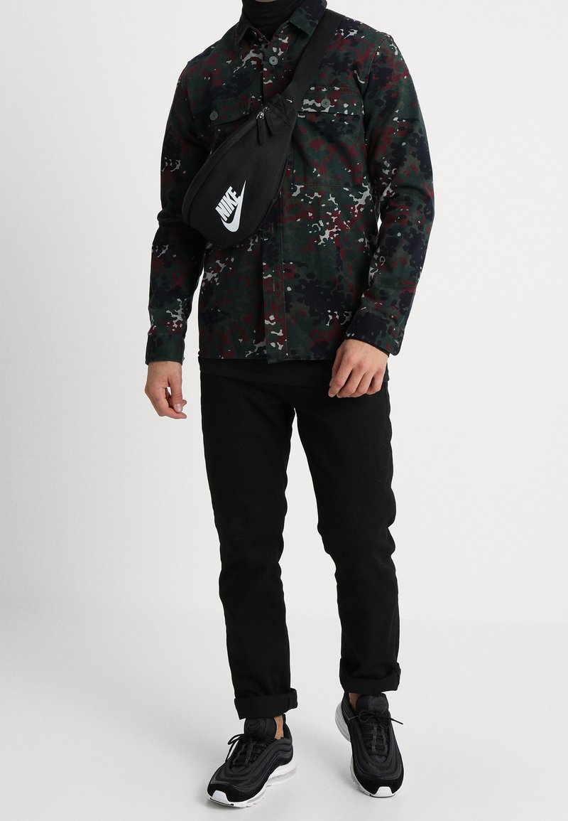 Homme portant une veste camouflage foncée, un pantalon noir, un sac bandoulière Nike noir et des baskets noires avec semelles blanches sur un fond uni.