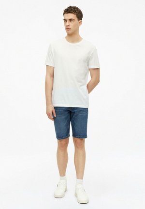 Junger Mann steht mit den Händen hinter dem Rücken, trägt ein schlichtes weißes T-Shirt, blaue Jeansshorts, weiße Socken und weiße Sneaker.