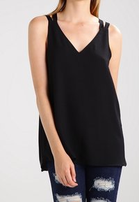 Top negro sin mangas con escote en V y tirantes finos; hecho de una tela suave con un corte holgado; combinado con unos jeans azules desgastados.