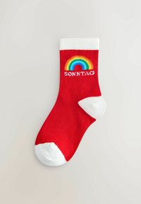 Calzino rosso alla caviglia con un polsino a costine bianche, una grafica arcobaleno e la parola "SONNTAG" in bianco sopra l'arcobaleno.