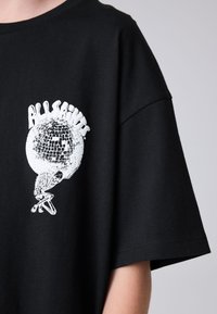 Fekete pamut póló, melyen fehér grafikai nyomat egy csontvázról, amely egy diszkógömböt tart, környakú nyakkivágással és rövid ujjakkal.