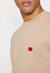 Gros plan d'une personne portant un pull en maille beige avec un petit logo carré rouge portant la mention « HUGO » sur la poitrine.