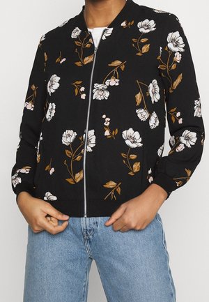 Schwarze Reißverschlussjacke mit weißem und braunem Blumenmuster, getragen über einem weißen Hemd und kombiniert mit hellblauen Jeans.