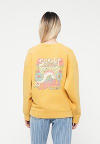 Gul genser med en fargerik grafisk trykk på baksiden som viser blomster- og bølgedesign, ribbede mansjetter og rund hals.