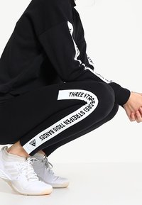 Leggings noirs avec du texte blanc sur le côté indiquant "THREE STRIPES TIES BANDS", associés à un sweat-shirt noir et des chaussures de sport blanches.