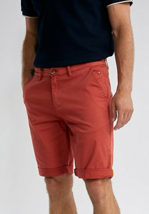 Homme portant un short rouille retroussé et une chemise à manches courtes bleu marine, debout les mains près des poches.