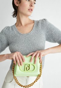Borsa verde menta con una finitura testurizzata, tracolla in catena dorata e un design distintivo con chiusura circolare. Modella che indossa un top lavorato a maglia argento.