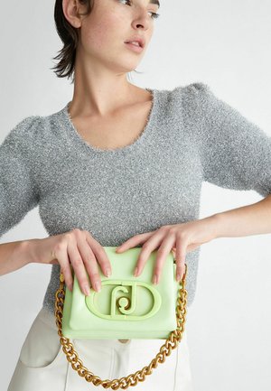 Borsa verde menta con una finitura testurizzata, tracolla in catena dorata e un design distintivo con chiusura circolare. Modella che indossa un top lavorato a maglia argento.