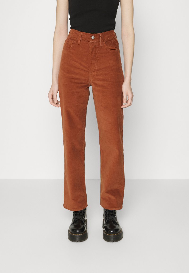 Frau trägt hochgeschnittene, rostfarbene Cordhosen, ein schwarzes, eng anliegendes ärmelloses Oberteil und schwarze Schnür-Plateau-Stiefel.