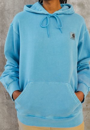 Personne portant un sweat à capuche Carhartt bleu clair avec les mains dans la poche avant devant un mur gris texturé.