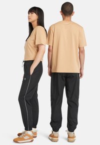 Beige katoenen t-shirt met een ronde hals, gecombineerd met zwarte joggerbroeken met zijzakken en witte piping, gedragen met bruine wandelschoenen.