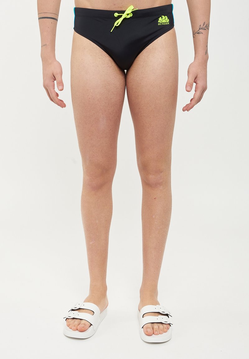 Slip da bagno neri con cordino verde e dettagli blu. Indossati con sandali bianchi, che mettono in risalto una trama di tessuto liscia e un design aderente.