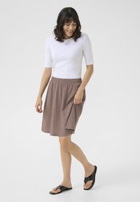 Jupe brune légère avec une taille élastique, accompagnée d'une chemise blanche ajustée et de sandales noires ouvertes, portée dans une pose décontractée.