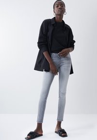 Schwarze Knopfleiste über einem schwarzen T-Shirt, kombiniert mit hellgrauen Skinny Jeans und schwarzen Slide-Sandalen. Einfaches Design mit lässiger Passform.