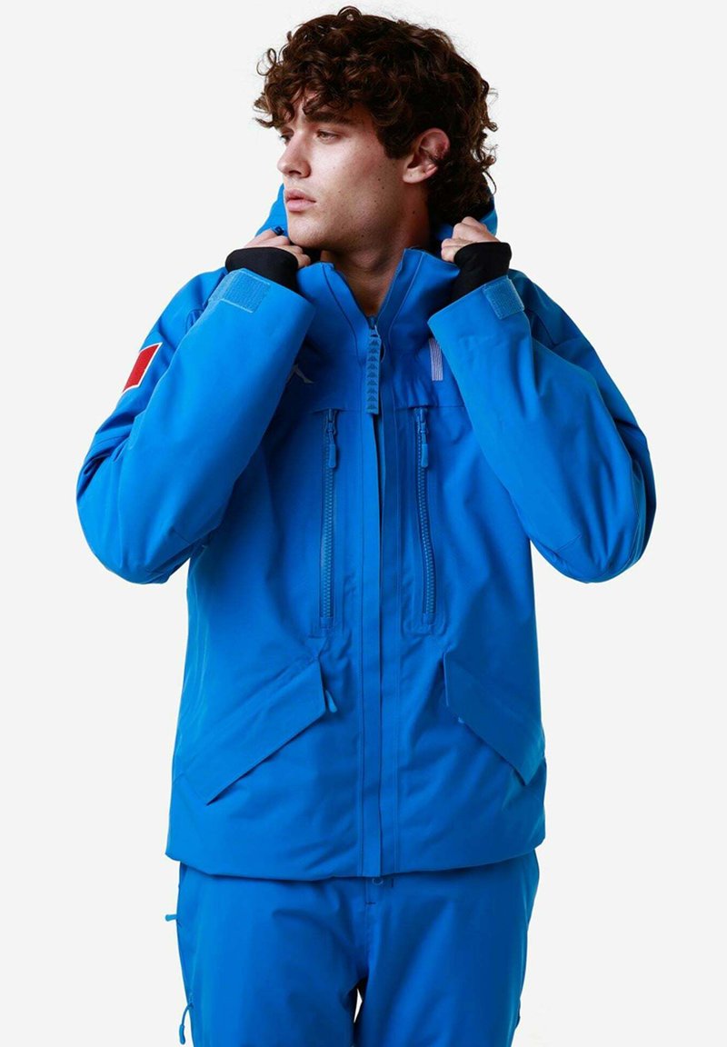 Giacca impermeabile blu con collo alto e cappuccio regolabile. Presenta due tasche frontali e un dettaglio del logo rosso a contrasto sulla manica.