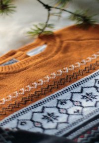 Pull en tricot orange avec un motif décoratif en blanc, noir et bleu, présentant des motifs géométriques et floraux le long de l'ourlet.