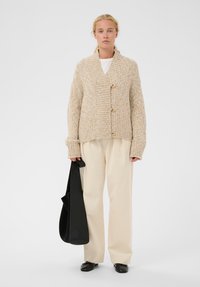 Frau trägt eine beige Strickjacke, ein weißes Oberteil, cremfarbene Pluderhosen, schwarze Ballerinas und hält eine große schwarze Tragetasche. Sie steht vor einem schlichten weißen Hintergrund.