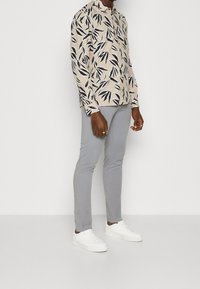 Long-sleeve shirt in beige with black leaf pattern, paired with light gray slim-fit pants and white sneakers. The outfit features a button-up design.