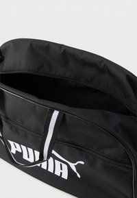 Μαύρη αθλητική τσάντα Puma με λευκό λογότυπο, μπροστινή τσέπη με φερμουάρ, κεντρικός χώρος ανοιχτός και μαύρη-λευκή ριγέ λαβή πάνω σε λευκό φόντο.