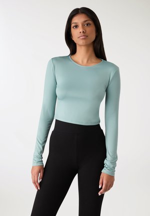 Długi rękaw, krótka bluzka w jasnym morskim odcieniu, wykonana z gładkiej, elastycznej tkaniny, z okrągłym dekoltem. W zestawie z czarnymi legginsami z wysokim stanem.