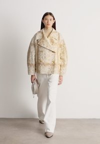 Sieviete staigā valkājot lielizmēra bēša rakstaina žaketi, krēmkrāsas augstu uzvilktu džemperi, baltas platām biksēm, bēšas kurpes un tur rokās somiņu, kas saskaņota ar apģērbu.