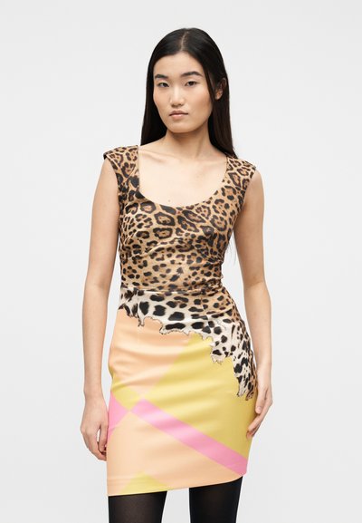 Νεαρή γυναίκα με μακριά σκούρα μαλλιά που φοράει αμάνικο φόρεμα με τοπ σε λεοπάρ σχέδιο και γεωμετρική φούστα σε κίτρινο, ροδάκινο και ροζ χρώμα.