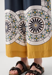 Flowy pants with a patterned fabric featuring blue, white, green, and yellow hues; solid yellow hem; paired with black sandals.