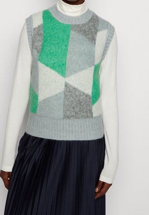 Modell trägt ärmellose Strickweste mit grünen, grauen und weißen geometrischen Mustern über weißem Rollkragenpullover und schwarzem, plissiertem Rock.