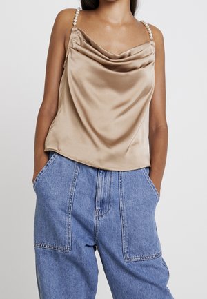 Kvinde iført en beige satin camisole med perlespænder og højtaljede løse blå denimjeans med hænderne i lommerne.