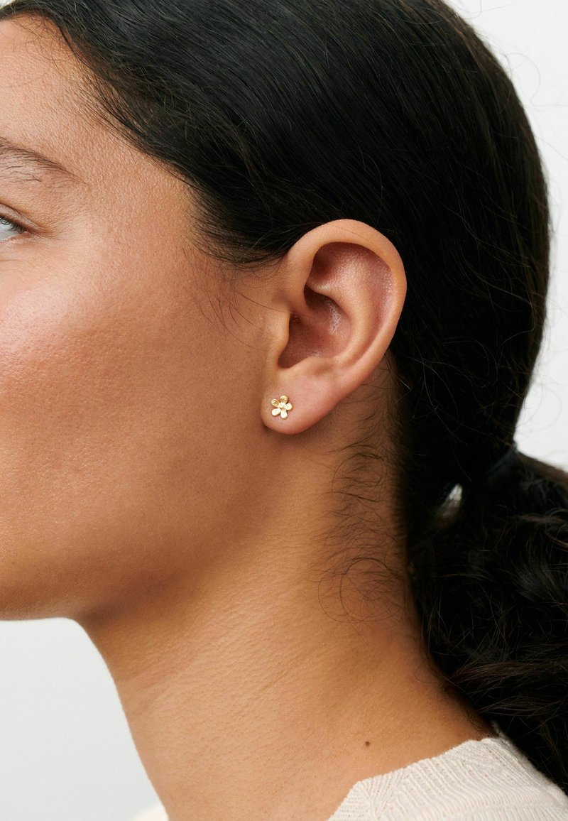 Gros plan sur l'oreille gauche d'une femme portant une petite boucle d'oreille en forme de fleur en or, les cheveux foncés tirés en arrière en une queue de cheval basse.