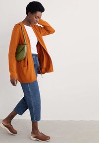 Orange knitted cardigan with pockets, white t-shirt, blue denim cropped pants, and brown wooden clogs. Green handbag slung over the shoulder.